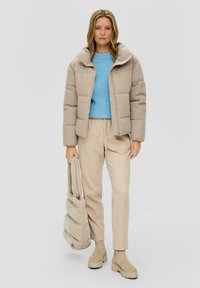 Beige Puffer-Jacke mit gestepptem Design, Reißverschluss und Kapuze. Hellblaues Strickoberteil darunter. Beigefarbene lockere Hose und Tasche. Braune Stiefeletten.