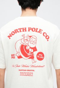 Hvid bomulds-T-shirt med en rød grafik af Julemanden med en sæk, tekst i rødt og accenter med ordene "NORTH POLE CO." og "OPEN TIL LATE."
