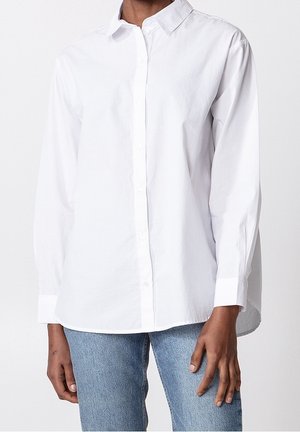 Personne portant une chemise blanche simple à manches longues boutonnée et un jean bleu clair, posant devant un fond blanc uni.