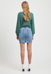 Pull vert en maille avec des manches bouffantes associé à une mini-jupe en denim bleu clair. Modèle portant des bottesblanches à la cheville. Vue arrière.