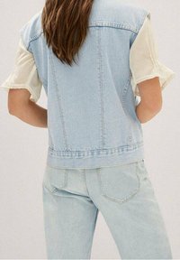Helle blaue Denim-Weste mit lockerer Passform, rohem Saum und unfertigen Ärmeln, über einem weißen, kurzärmligen Oberteil getragen. Dazu helle Jeans.