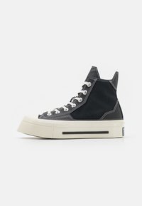 Zapatilla alta con parte superior de lona negra, costuras blancas en contraste y suela gruesa de goma crema. Presenta cordones redondos y un reverso texturizado.