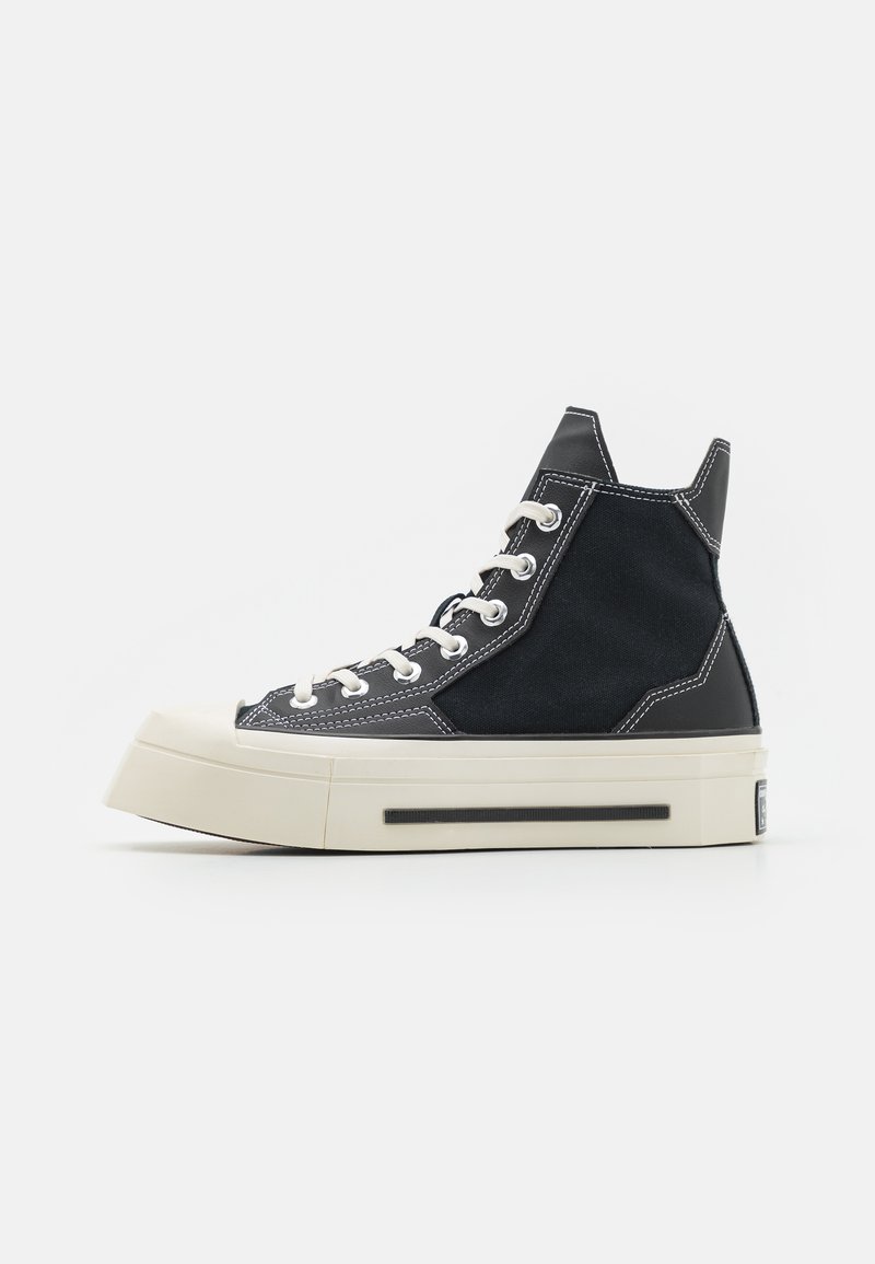 Zapatilla alta con parte superior de lona negra, costuras blancas en contraste y suela gruesa de goma crema. Presenta cordones redondos y un reverso texturizado.