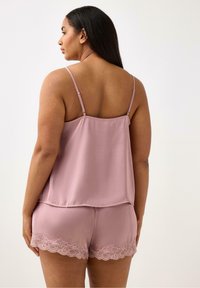 Vaaleanpunainen satiininen pyjama-asusetti, johon kuuluu olkaimeton paita, jossa on vauhdikas muotoilu, ja pitsireunaiset shortsit. Silkkinen pinta ja hienovaraiset yksityiskohdat.