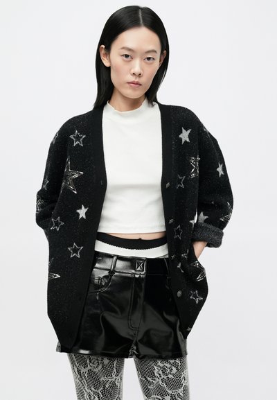 Cardigan negru tricotat cu modele de stele argintii, închidere cu nasturi, croială oversized. Asortat cu un top alb, pantaloni scurți negri lucioși și dresuri imprimate.