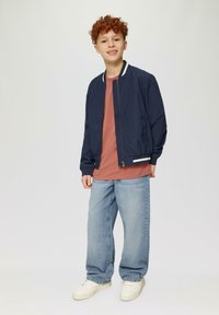 Marine bomberjack met witte afwerking, gecombineerd met een lichtroze T-shirt en lichtblauwe wijde jeans. Draagt witte sneakers.