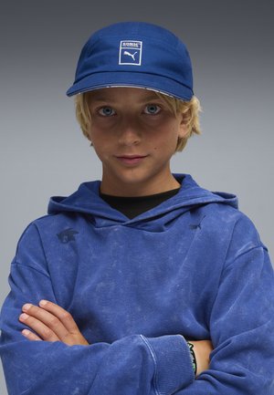 Gorra azul con un parche de logo blanco, combinada con una sudadera azul que presenta un logo sutil. La tela muestra una apariencia texturizada y desgastada.