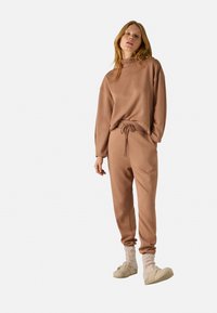 Sudadera marrón con cuello alto y corte relajado, combinada con pantalones de chándal a juego que cuentan con un cordón ajustable en la cintura.