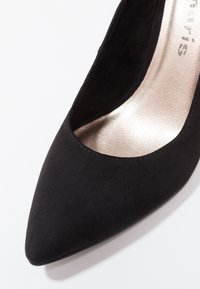 Escarpin à talon haut en daim noir à bout pointu avec une semelle intérieure métallique claire et une texture lisse sur fond blanc.
