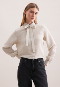 Eine Frau trägt eine cremefarbene Bluse mit feinen Streifen und einem Kragen mit Schleife sowie hochgeschnittene schwarze Jeans. Sie steht mit den Händen in die Hüften gestützt vor einem schlichten Hintergrund.