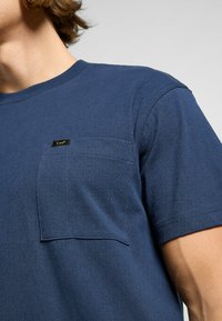 Sininen puuvillapaita, jossa on pyöreä pääntie, lyhyet hihat ja vasemmalla rintapuolella tasku. Lisäksi paidassa on pieni musta etiketti, jossa lukee "Lee."