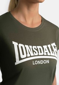 Olive green t-shirt with a round neckline and short sleeves. Features bold white "LONSDALE LONDON" lettering across the chest. Soft fabric.