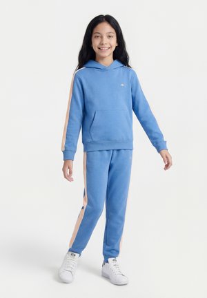 Κορίτσι που χαμογελά, όρθιο, φορώντας μπλε φούτερ με κουκούλα και αντίστοιχο παντελόνι με ριγέ ροδάκινου χρώματος, καθώς και λευκά αθλητικά παπούτσια σε λευκό φόντο.