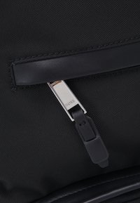 Sac en tissu noir avec une sangle en cuir lisse et une tirette de fermeture éclair en argent avec un détail de logo. Surface extérieure texturée avec des coutures visibles.