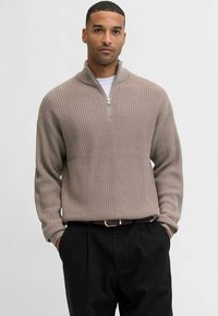 Suéter de punto tejido de color marrón claro con cuello de media cremallera, combinado con pantalones negros; cuenta con mangas largas y un corte relajado.