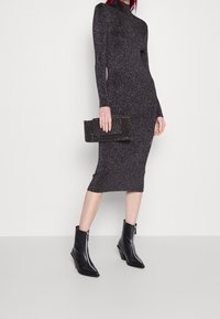 Robe noire côtelée à manches longues avec un col montant, assortie à une pochette noire texturée et des bottines, avec un détail zippé.