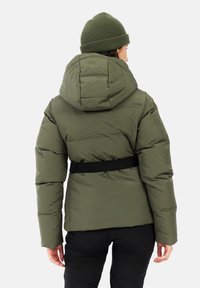 Olijfgroene pufferjack met een capuchon, ceintuurtje in de taille en een doorgestikte textuur. Gecombineerd met een bijpassende gebreide muts. Zwarte broek zichtbaar.