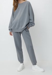 Szara bawełniana bluza z opadającymi ramionami w zestawie z dopasowanymi dresami. Strój cechuje luźny krój i ściągacze przy rękawach, uzupełnione białymi sportowymi butami.