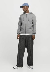 Graue Zip-Hoodie mit Kordelzügen, kombiniert mit lockeren dunklen Jeans. Eine hellblaue Kappe rundet das Outfit ab, und graue Sportschuhe vervollständigen den Look.