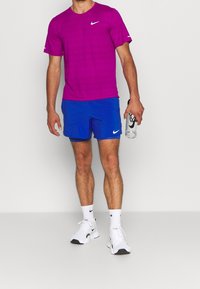 Hombre con camiseta púrpura de Nike, pantalones cortos azules, calcetines blancos y zapatillas blancas, sosteniendo una botella de agua transparente de Nike.