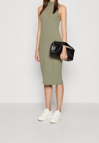 Robe midi sans manches à côtes de couleur vert olive avec un col montant, associée à une pochette noire et des baskets blanches. Texture lisse et design ajusté.