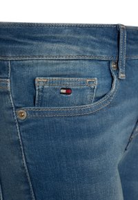 Vaqueros de mezclilla azul claro con un bolsillo trasero que presenta un pequeño logo bordado en rojo, blanco y azul. La tela muestra una textura ligeramente desgastada.