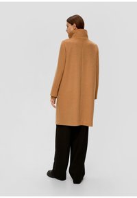 Manteau en laine couleur chameau avec un col montant, coupe droite et manches longues, associé à un pantalon large noir et des chaussures plates.