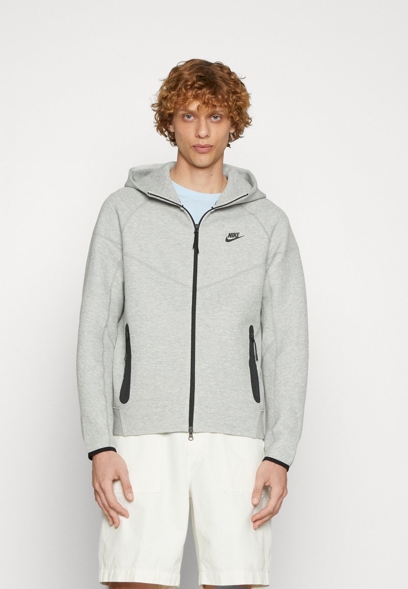 Sudadera gris de Nike con cierre frontal, acentos en negro y bolsillos laterales. Presenta una capucha y una textura suave. Diseño ligero para uso casual.