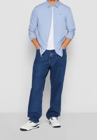 Ljusblå skjorta, vit t-shirt under, blå denimbyxor och vita sneakers med mörka accenter och strukturerade sulor.