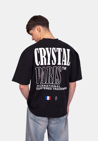 T-shirt oversized noir avec un grand texte blanc "CRYSTAL PARIS" au dos, comprenant un petit drapeau français et un détail logo en forme de diamant.