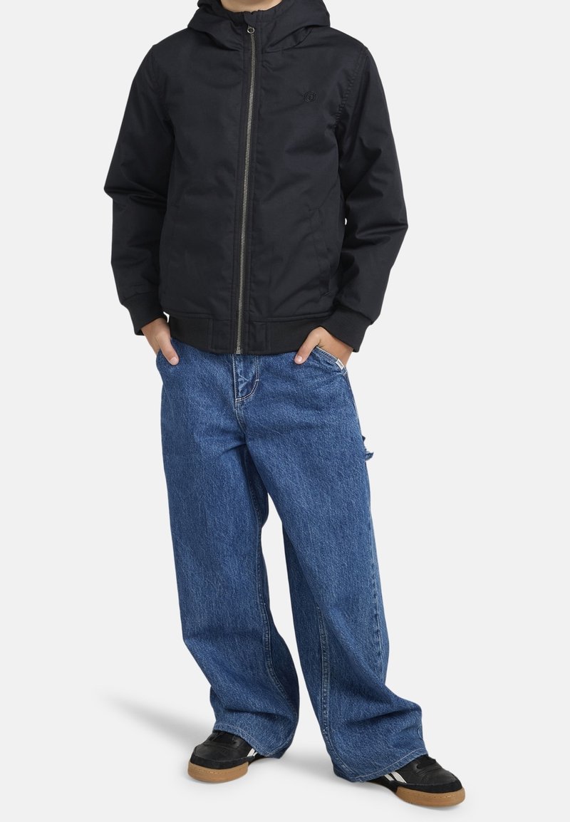 Jeune personne portant une veste à capuche noire, un jean bleu large et des baskets noires avec des semelles en gomme, les mains dans les poches, debout sur un fond blanc.