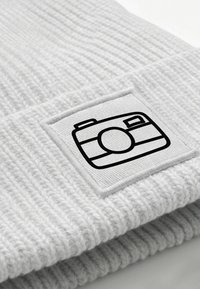 White ribbed knit beanie with a square woven patch featuring a black camera icon. Soft texture, casual design.