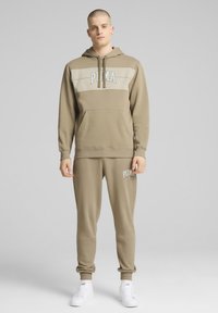 Felpa con cappuccio color tan con una striscia beige chiaro che presenta un logo, abbinata a pantaloni jogger coordinati. Design semplice, tasca frontale e polsini a coste.
