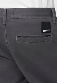 Pilkos džinsinės kelnes su lygia tekstūra, su užpakaline kišenė ir kvadratine juoda etikete su balta tekstu. Tvarkingas siuvimas ir modernaus kirpimo.