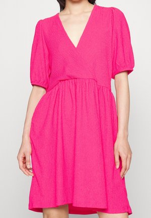 Frau trägt ein leuchtend pinkfarbenes, strukturiertes Kleid mit Puffärmeln und V-Ausschnitt, steht vor einem schlichten hellen Hintergrund.