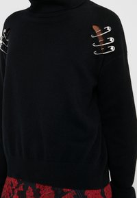 Schwarzer Pullover mit Rollkragen, Ausschnitten an den Schultern und vier silbernen Sicherheitsnadeln als Akzenten. Weicher, gestrickter Stoff mit geripptem Saum.