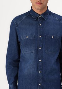 Camisa de деним en azul oscuro, con cuello abotonado, bolsillos delanteros con cierres a presión y mangas largas con puños abotonados.