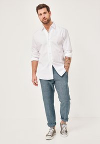White long-sleeve button-up shirt, light blue textured pants, and beige sneakers with white laces. Features rolled-up sleeves and visible tattoos.