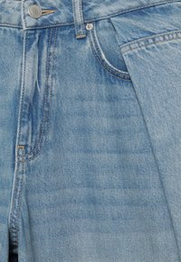 Blugii din denim albastru deschis văzuți de aproape, evidențiind butonul frontal, bucla pentru curea și buzunarul cu cusături contrastante portocalii.