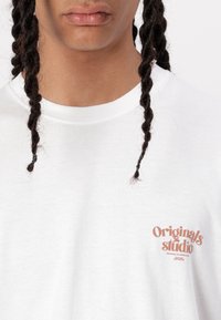 White cotton t-shirt with a crew neckline and short sleeves. Features a small, printed "Originals Studio" logo in an orange-brown color.