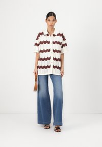 Camicia a maniche lunghe in maglia con strisce ondulate marroni su sfondo bianco sporco, abbinata a jeans a gamba larga blu chiaro e sandali neri con fasce.