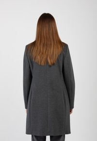 Donna con lunghi capelli castani lisci, che indossa un cappotto grigio scuro, in piedi di spalle contro uno sfondo grigio chiaro e uniforme.