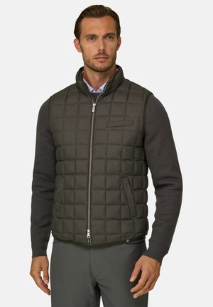 Homme portant un gilet matelassé foncé zippé par-dessus une chemise en tricot à manches longues avec un pantalon foncé assorti, debout et regardant devant lui.