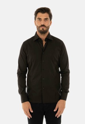 Uomo con capelli castani e barba che indossa una camicia nera aderente a maniche lunghe con bottoni, in piedi davanti a uno sfondo bianco semplice.