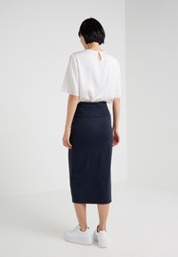 Witte top met korte mouwen en een sleutelgatdetail aan de achterkant, gecombineerd met een navy kokerrok, gedragen met witte sneakers op een effen achtergrond.