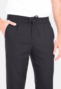Pantaloni neri con vita regolabile tramite cordoncino, dotati di chiusura a bottone e pieghe frontali. Il tessuto appare liscio e leggero.