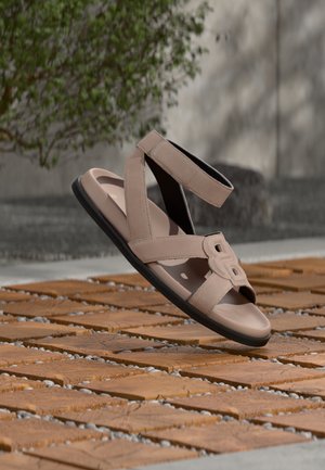 Sandale beige avec un design à lanières croisées, tige texturée et semelle plate noire, ornée d'une boucle décorative à l'avant.