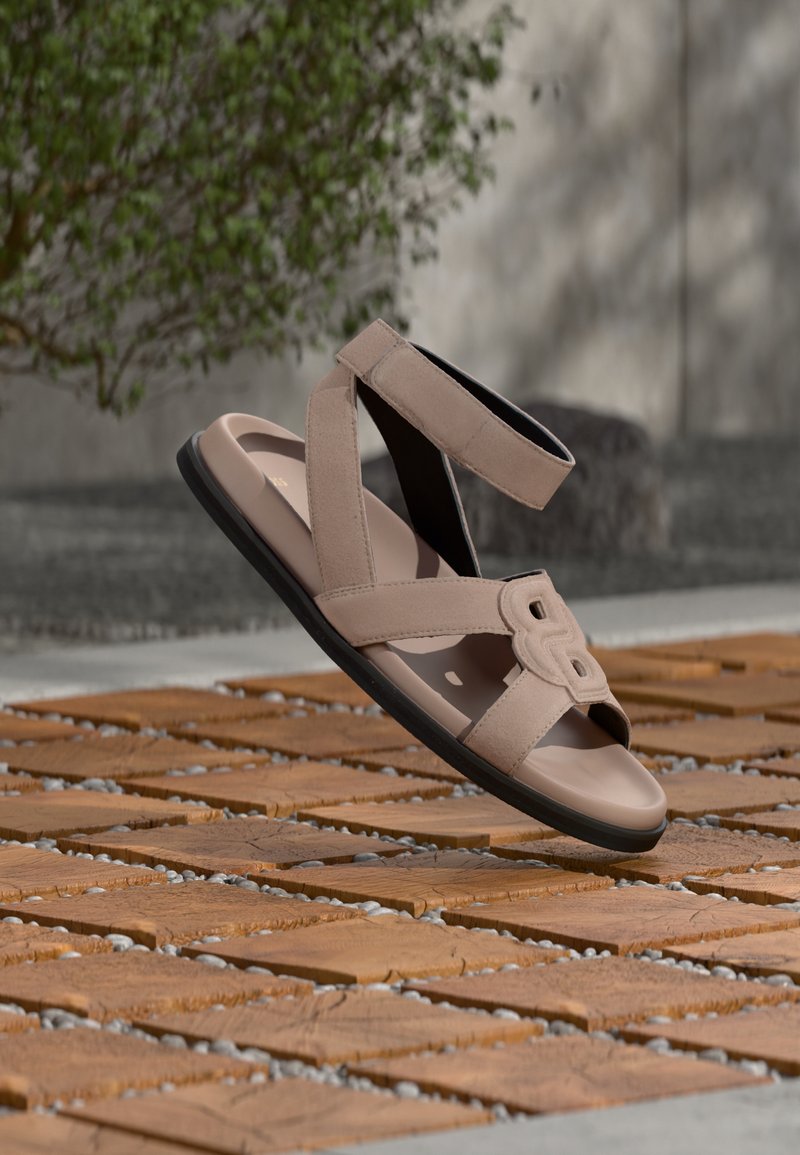 Μπεζ σανδάλι με σχεδιασμό σταυρωτού λουριού, υφή στην επάνω πλευρά και μαύρη επίπεδη σόλα, με διακοσμητικό αγκράφα στο μπροστινό μέρος.