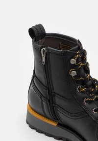 Black leather boot with a zip closure, yellow-accented laces, padded collar, and textured rubber sole. Features stitching details and pull tab.