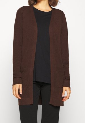 Frau trägt eine dunkelbraune, offene Strickjacke mit langen Ärmeln und Fronttaschen über einem schwarzen Hemd und schwarzen Hosen.